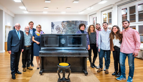 Eine Aufnahme, die bei der Ausstellungseröffnung in den Kunstsammlungen Chemnitz entstand. In der Bildmitte befindet sich das Klavier der jüdischen Familie Margulies in dem Ausstellungsraum auf einem sandfarbenen Teppich, der wiederum auf dem hellen Parkett des Ausstellungsraumes platziert ist. Vor dem Klavier steht ein schwarzer, geschwungener Klavierhocker aus Holz, die runde Sitzfläche ist aus einem hellen Material. Dahinter steht das Klavier. Es hat die gleiche schwarze Farbe wie der Hocker. Die Tastenklappe ist geschlossen. Die dahinterliegende Fläche des Klaviers ist mit Prägungen versehen – darunter eine, die sich mittig in einem Achteck befindet und ein florales Ornament aufweist. Rechts und links vom Klavier stehen Oberbürgermeister Sven Schulze gemeinsam mit der Sächsischen Staatsministerin Barbara Klepsch und der Familie Margulies. Alle lächeln und schauen in die Kamera. Im Hintergrund ist eine Fotowand erkennbar, die einen belaubten Baum zeigt. Der Ausstellungsraum besitzt weiße Wände. Hinter den Fenstern auf der rechten Seite ist es dunkel; von oben werfen Lampen weißes Licht auf die Szene.