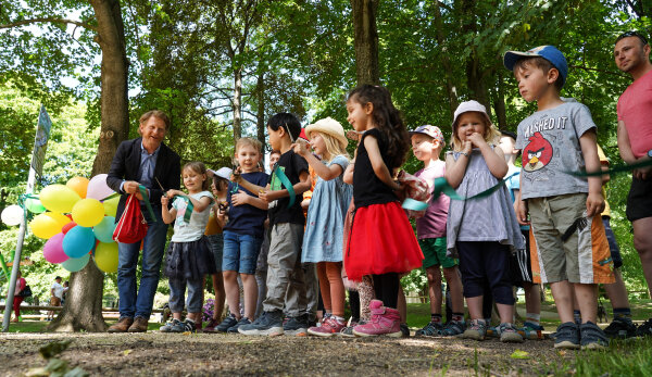 Rechts im Bild sind Kinder bei der Einweihung des Schloßteich-Spielplatzes zu sehen. Links im Bild steht Bürgermeister Michael Stötzer neben bunten Luftballons. Vor den Kindern ist ein Band zu sehen, welches von einem Mädchen mit einer Schere zerschnitten wird.