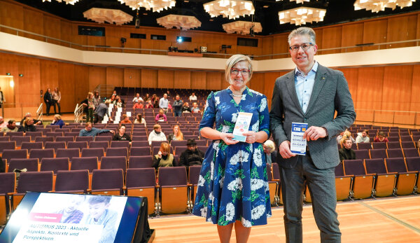 Im Vordergrund präsentieren Frau Liebetrau, die Behindertenbeauftragte der Stadt Chemnitz und Bürgermeister Ralph Burghart die Programm-Flyer zur ersten Chemnitzer Autismuswoche. Im Hintergrund ist der Veranstaltungssaal des Carlowitz-Congresscenters zu sehen.