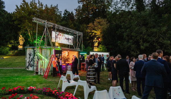 Im Vordergrund des Bildes sind weiße Plastikstühle, Blumen und zahlreiche Personen erkennbar, die auf einer Wiese stehen. Links daneben befindet sich ein einzelstehendes Garagengebäude mit bunten Lichtern. Im Hintergrund ist eine parkähnliche Landschaft mit einer Wiese, Statuen und grün belaubten Bäumen erkennbar. Es herrscht Dämmerungslicht.