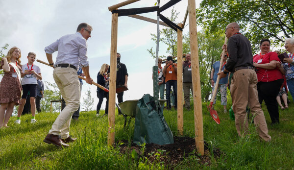 Bürgermeister Ralph Burghart legt an einem Apfelbaum weitere Erde mit einer roten Schaufel nach. Er trägt eine beigefarbene Hose und ein weißes Hemd. Neben ihm steht der Weimarer Bürgermeister Ralf Kirsten, bekleidet mit einem braunen Pullover und einer beigefarbenen Hose. Er hält ebenfalls eine Schaufel in den Händen. Im Hintergrund schauen einige Menschen zu. Die Sonne scheint.