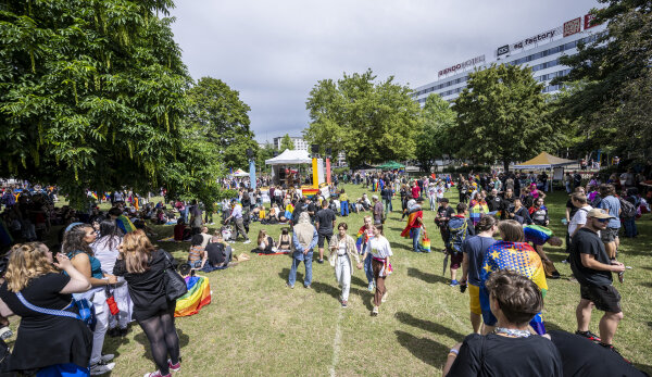 Auf der Wiese im Stadthallenpark sind zahlreiche Menschen versammelt. Viele von ihnen tragen Flaggen in Regenbogen-Farben. Auf der Wiese stehen vereinzelt Festzelte. Außerdem sind grün belaubte Bäume zu sehen. Rechts im Hintergrund ist das Rawema-Gebäude erkennbar. Die Sonne scheint.