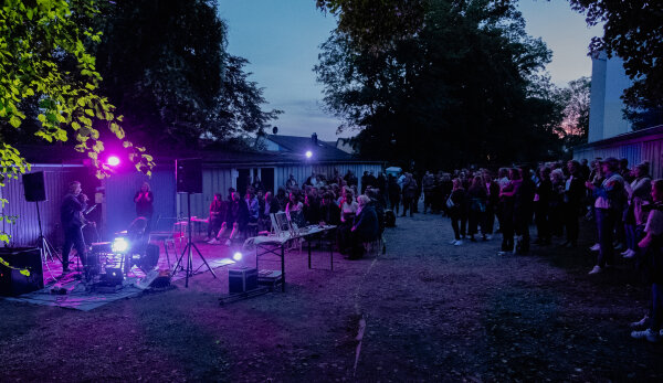 Links im Bild stehen vor einer Garagenreihe Musiker mit Licht, Musikanlage und Lautsprechern. Dahinter sitzen auf Bänken zahlreiche Zuhörende vor den Garagen. Es herrscht Abenddämmerung. 