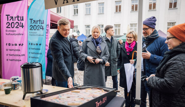 Kulturbürgermeisterin Dagmar Ruscheinsky steht mit Erni Kask vom Team Tartu2024 und Sachsens Kultur- und Tourismusministerin Barbara Klepsch auf einer Straße an einem Stand mit Tassen und einem Thermobehälter für Getränke. Erni Kask zeigt mit der Hand auf etwas. Alle tragen Mäntel. Links im Bild sind mehrere Banner in Blau und Pink zu sehen, auf denen „Tartu 2024“ steht. Im Hintergrund erkennt man eine gepflasterte Straße und ein Gebäude. Das Wetter ist schön.