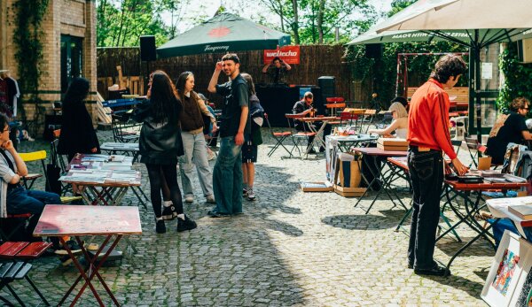 Das Bild zeigt den Innenhof des Weltechos, Tische und Stühle sind zu sehen. Leute sitzen auf den Stühlen oder stehen zwischen den Tischen auf dem Hof und unterhalten sich. Die Sonne scheint.