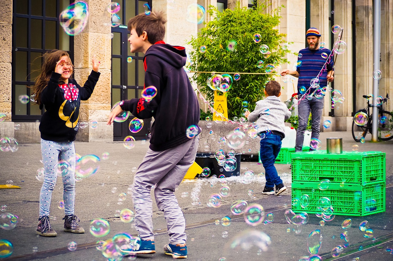 Drei Kinder spielen auf der Straße, viele Seifenblasen schweben durch die Luft, ein Erwachsener ist im Hintergrund zu sehen